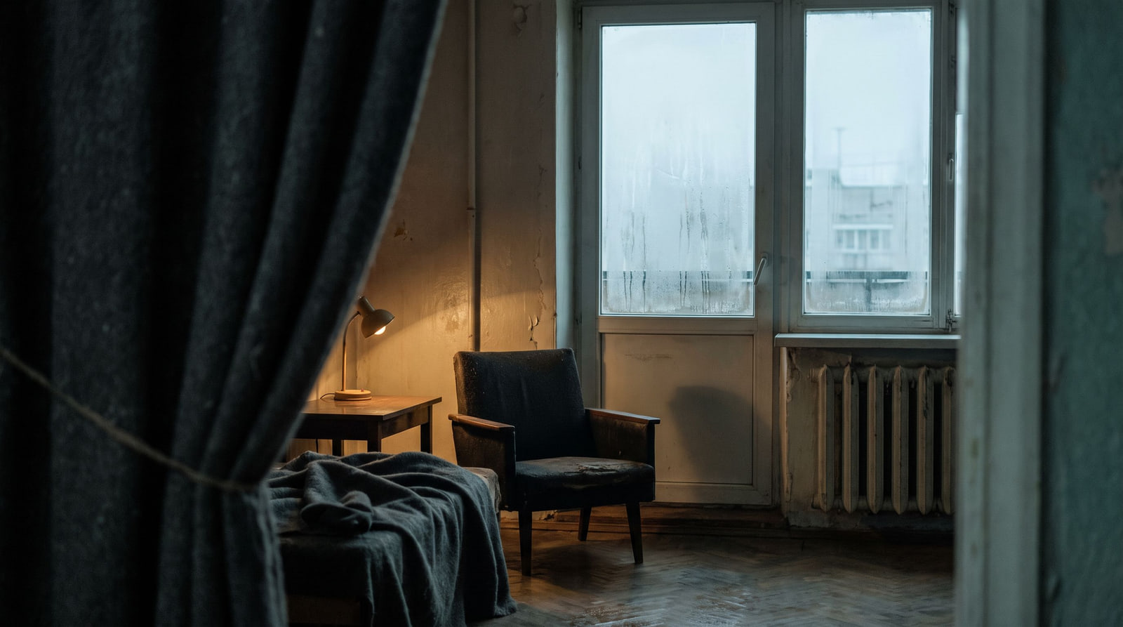 Dim lived-in room with dark textiles, a weak amber lamp and a cold fogged window.